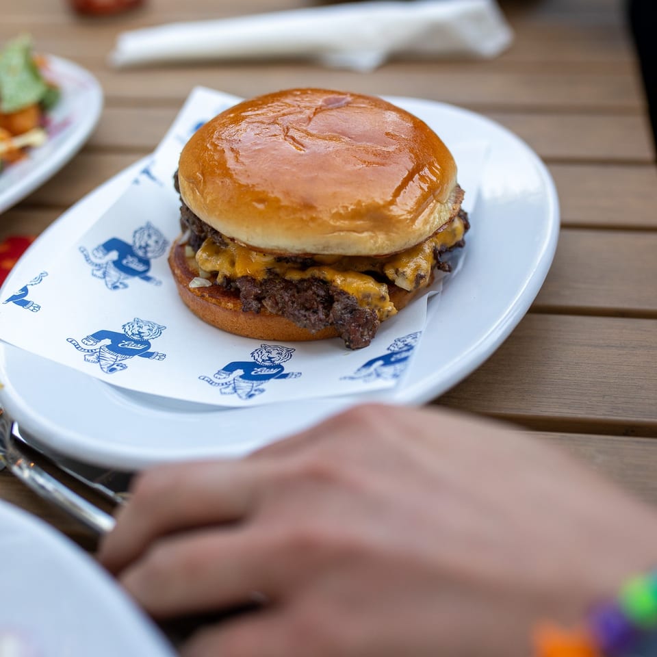 Double cheeseburger on a branded paper-lined plate at a wooden restaurant table, with diners and side dishes in the background.