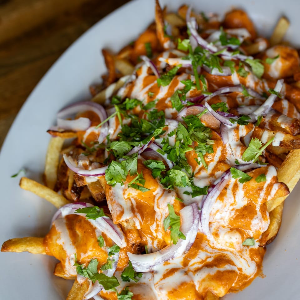 Loaded fries topped with orange and white sauces, red onion, and fresh cilantro on a white plate.