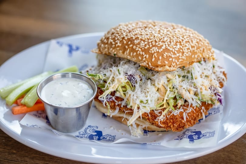 Crispy chicken sandwich with coleslaw, sesame bun, side of ranch, celery, and carrots on a white plate.
