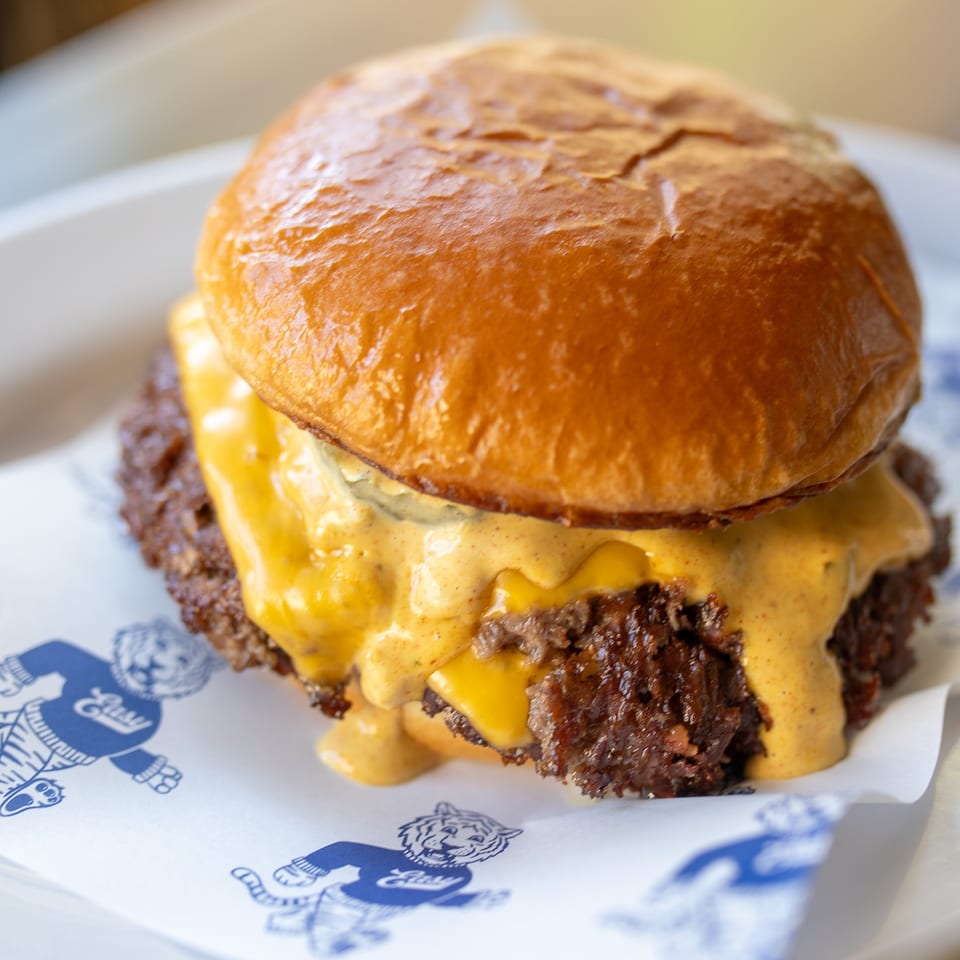 Close-up of a cheeseburger with a toasted bun, melted cheese, creamy sauce, and crispy beef topping on branded paper.