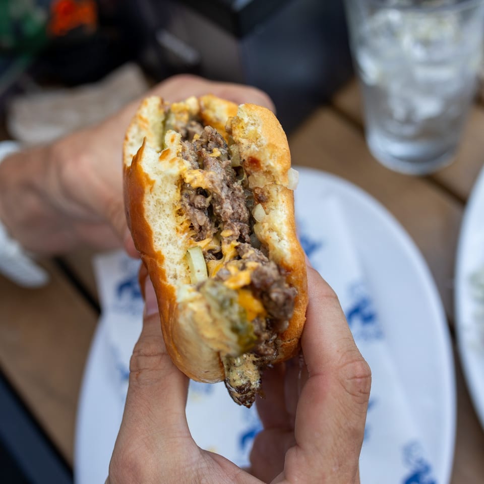 Hand holding a cheesesteak sandwich filled with sliced beef, melted cheese, and onions over a restaurant table.
